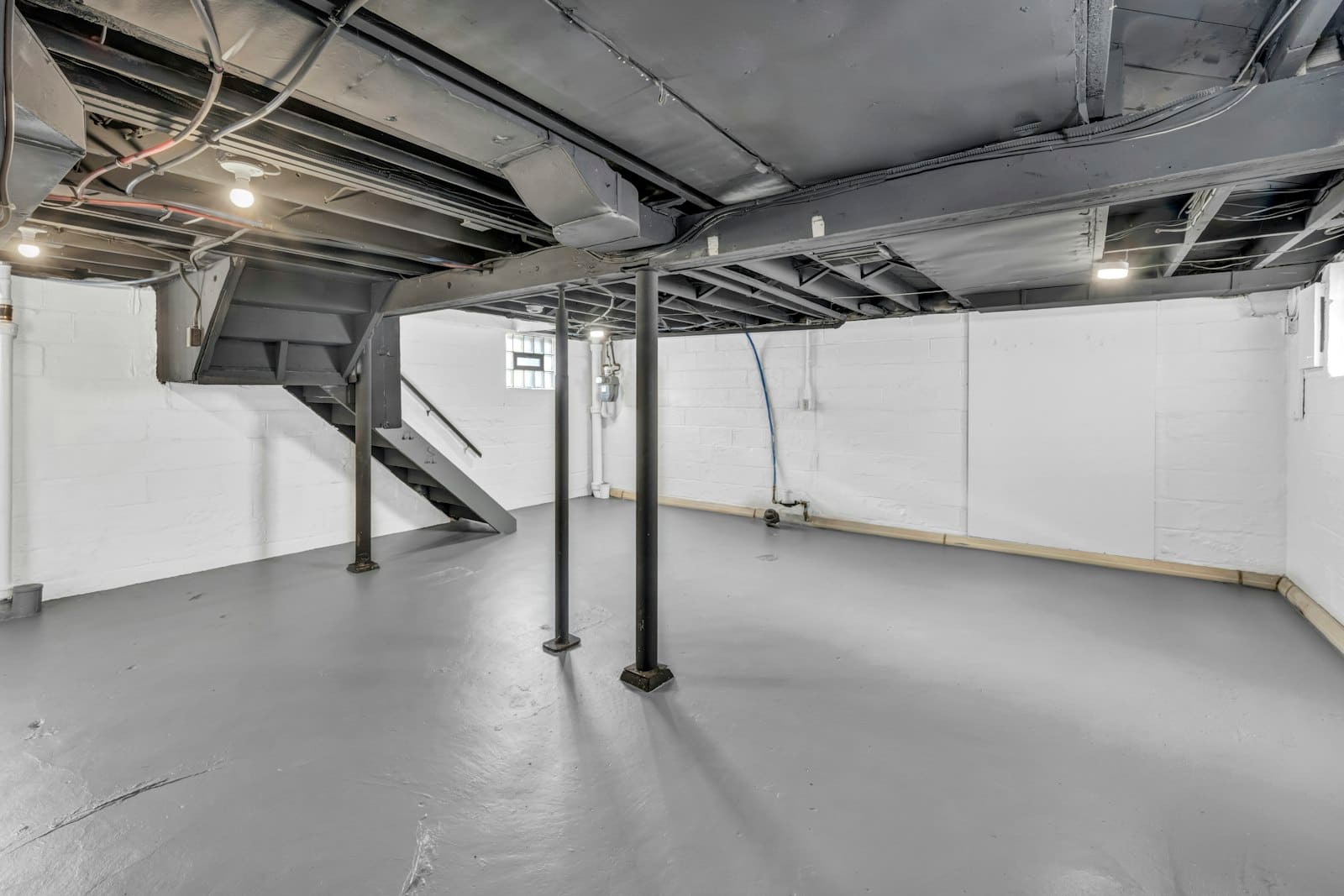 Sous-sol utilitaire vide au sol béton peint gris, poteaux métalliques et murs blancs, avant pose d'un revêtement de garage