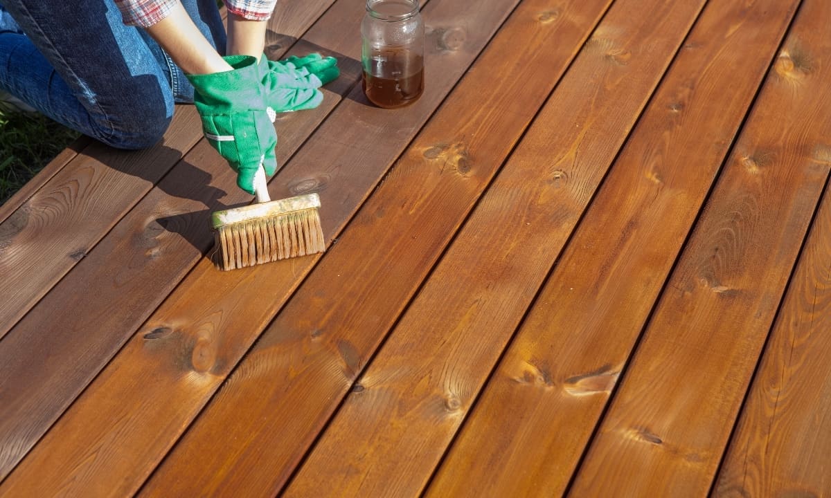 Application d'un saturateur à la brosse sur une terrasse en bois miel : les lames déjà traitées prennent une teinte plus chaude et satinée à côté du pot de produit.