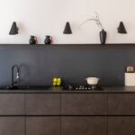 Voici une cuisine moderne et élégante avec un îlot central sombre, un évier noir, une plaque de cuisson et des luminaires noirs sur un mur blanc.