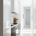 Une cuisine moderne, minimaliste, s'étend le long d'un couloir lumineux. Des armoires blanches et foncées abritent un four et une cuisinière. Le sol est clair et uniforme.