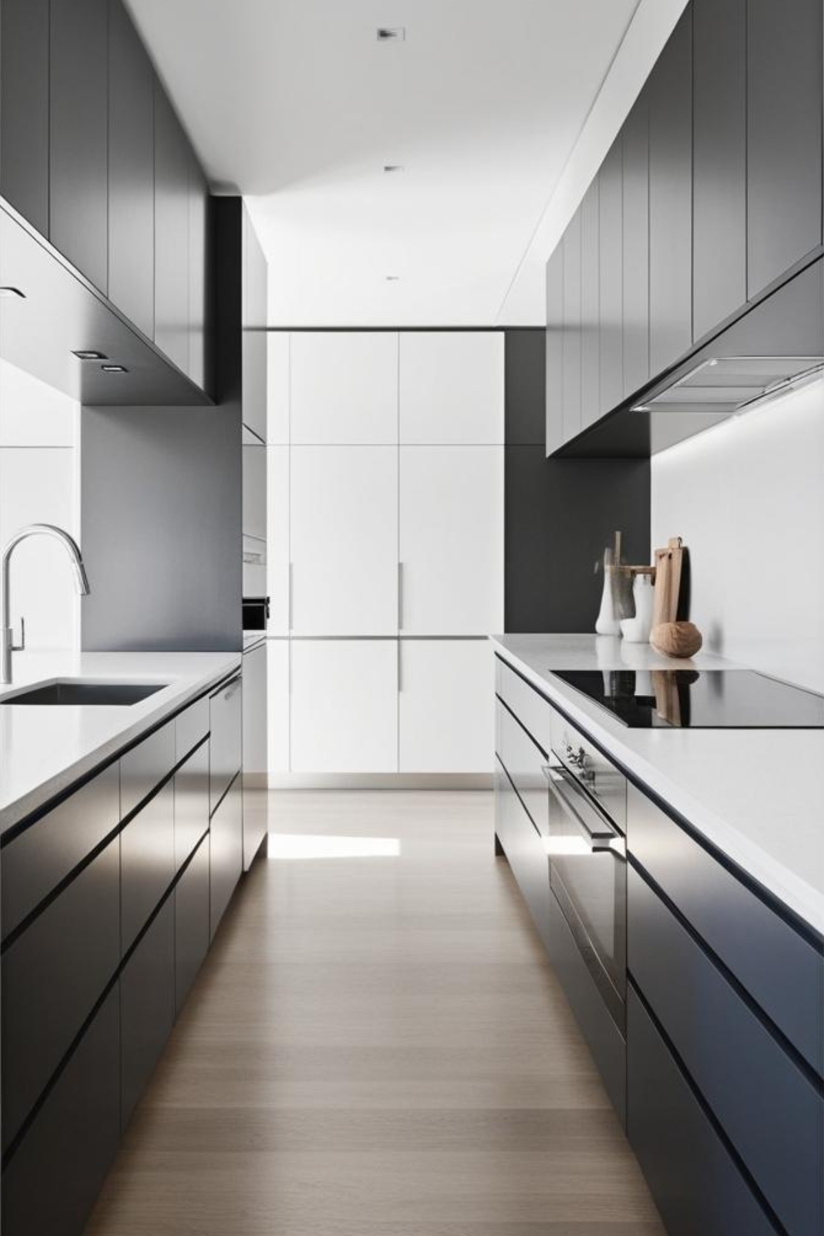 Une cuisine moderne est présentée. Des armoires noires et blanches sont disposées de chaque côté d'un sol clair. Un four encastré est visible. Le plan de travail est propre et épuré. L'espace est lumineux et minimaliste.