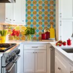 Une crédence en carrelage aux motifs circulaires bleus, oranges et verts orne une cuisine aux armoires blanches. Des bouquets de fleurs, des ustensiles de cuisine et des fruits sont disposés sur le comptoir. Le sol est en bois.