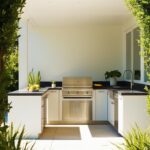 Un barbecue encastré en acier inoxydable est installé dans une cuisine extérieure moderne. Il est intégré à des armoires blanches, sous un auvent blanc, au milieu d'une végétation luxuriante.