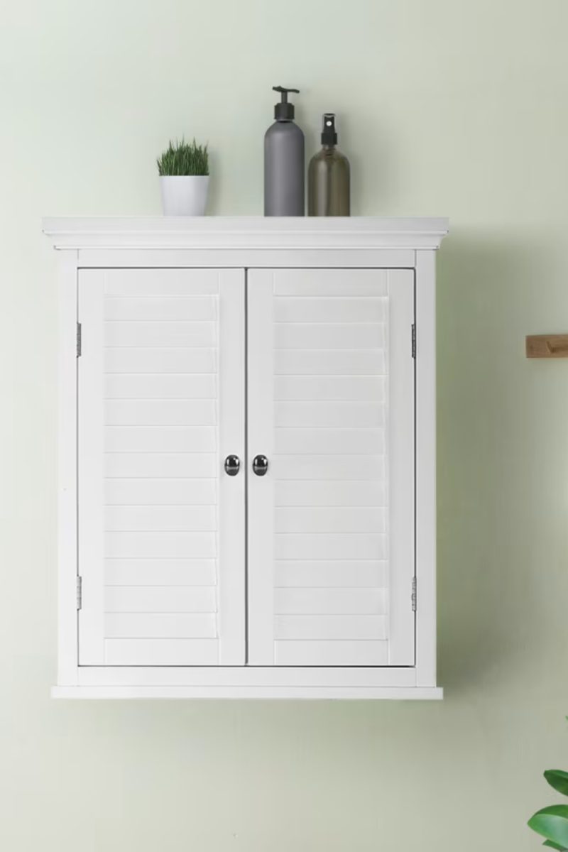 Voici une armoire blanche à persiennes accrochée au mur. Sur le dessus, on voit une plante en pot et deux bouteilles, une grise et une verte.
