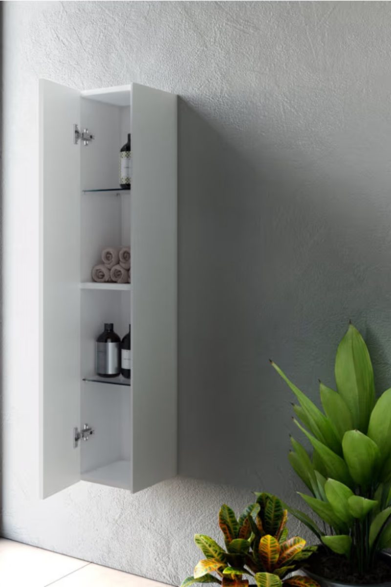 Un meuble de salle de bain blanc est fixé au mur avec une porte ouverte, révélant des étagères avec des serviettes et des bouteilles. Des plantes vertes sont au premier plan.