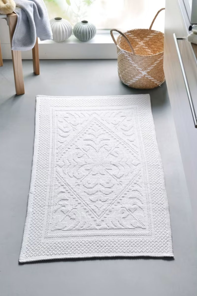 Un tapis de bain blanc à motifs est posé sur un sol gris, près d'une chaise en bois, d'un panier en osier et de vases décoratifs.