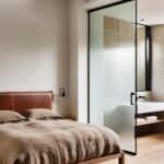 Une verrière d'atelier sépare la chambre à coucher de la salle de bains attenante, permettant à la lumière naturelle d'inonder l'espace. Un lit en bois massif avec une tête de lit en cuir et une couette en lin lavé beige invitent au repos. Le parquet en chêne massif et les poutres apparentes de la charpente renforcent le charme authentique de la pièce. Des touches de métal noir et de verre apportent une touche industrielle chic.