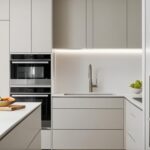 Une cuisine moderne et minimaliste. Des armoires et îlot couleur beige clair. Un four encastré est intégré. Un évier est situé dans un coin. Le tout est éclairé par des lumières encastrées.