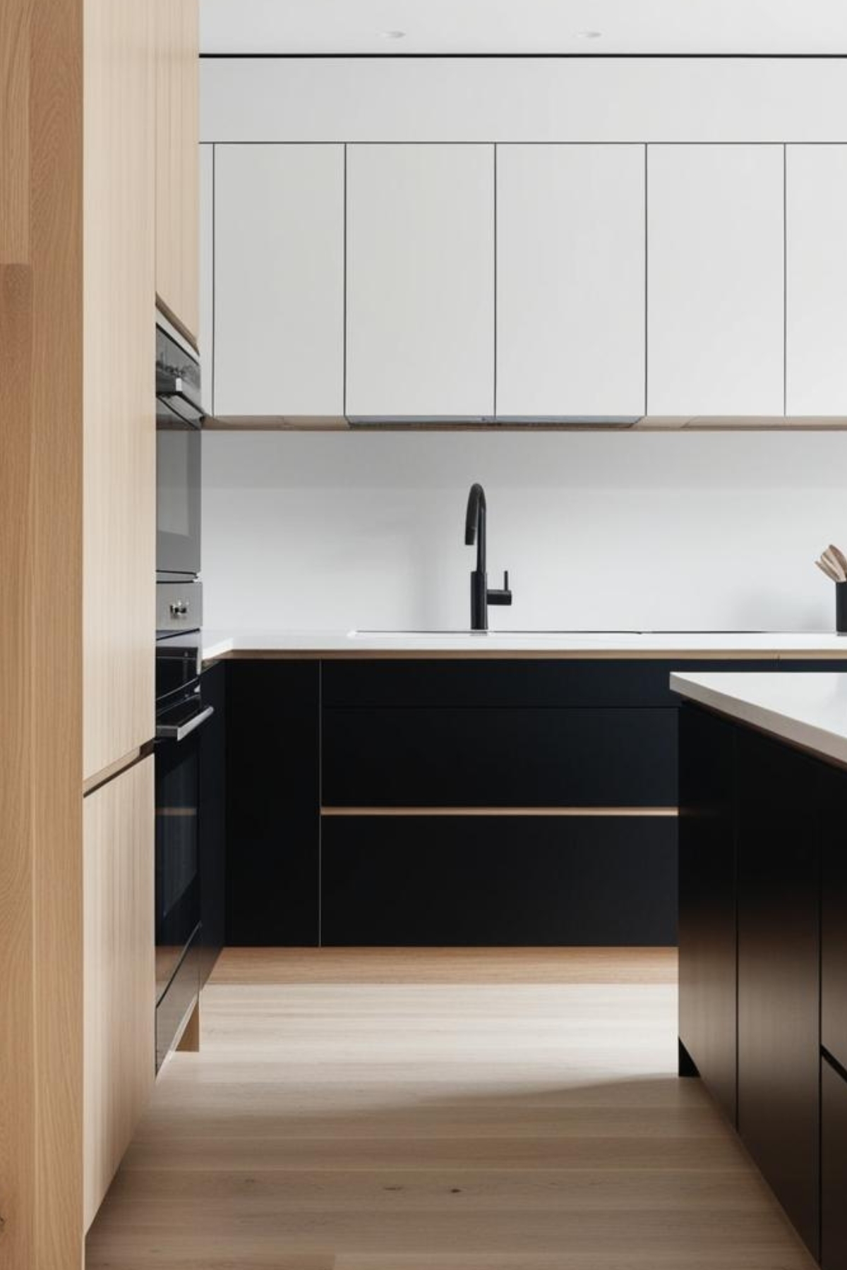 Une cuisine moderne est présentée. Des armoires blanches et noires se trouvent au-dessus et en dessous d’un évier avec robinet noir. Un four encastré est visible. Le sol est en bois clair.