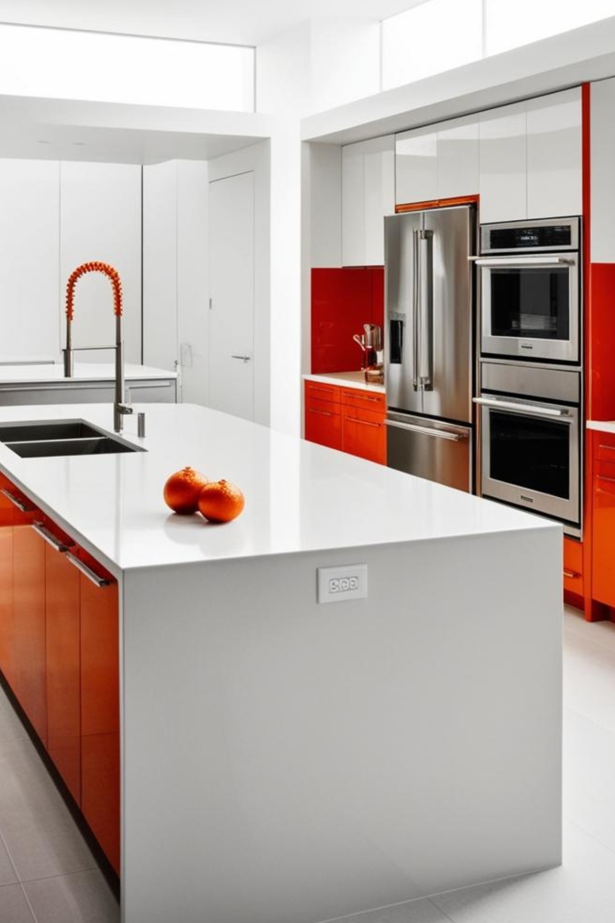 Une cuisine moderne blanche et orange vif. Une île blanche et orange domine la pièce, avec un évier et deux oranges dessus.