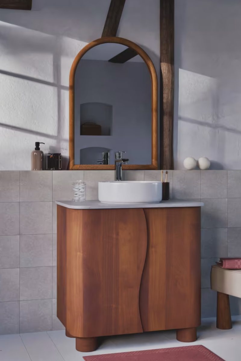 Meuble-lavabo en bois arrondi avec vasque blanche dans une salle de bain lumineuse. Un miroir en arc et un tabouret sont visibles, ajoutant à l'esthétique décorative.