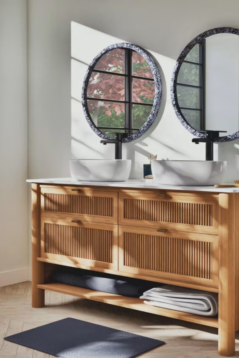 Un meuble-lavabo en bois clair supporte deux vasques blanches et des miroirs circulaires ornés. La pièce est lumineuse avec un tapis bleu sur un plancher en bois.