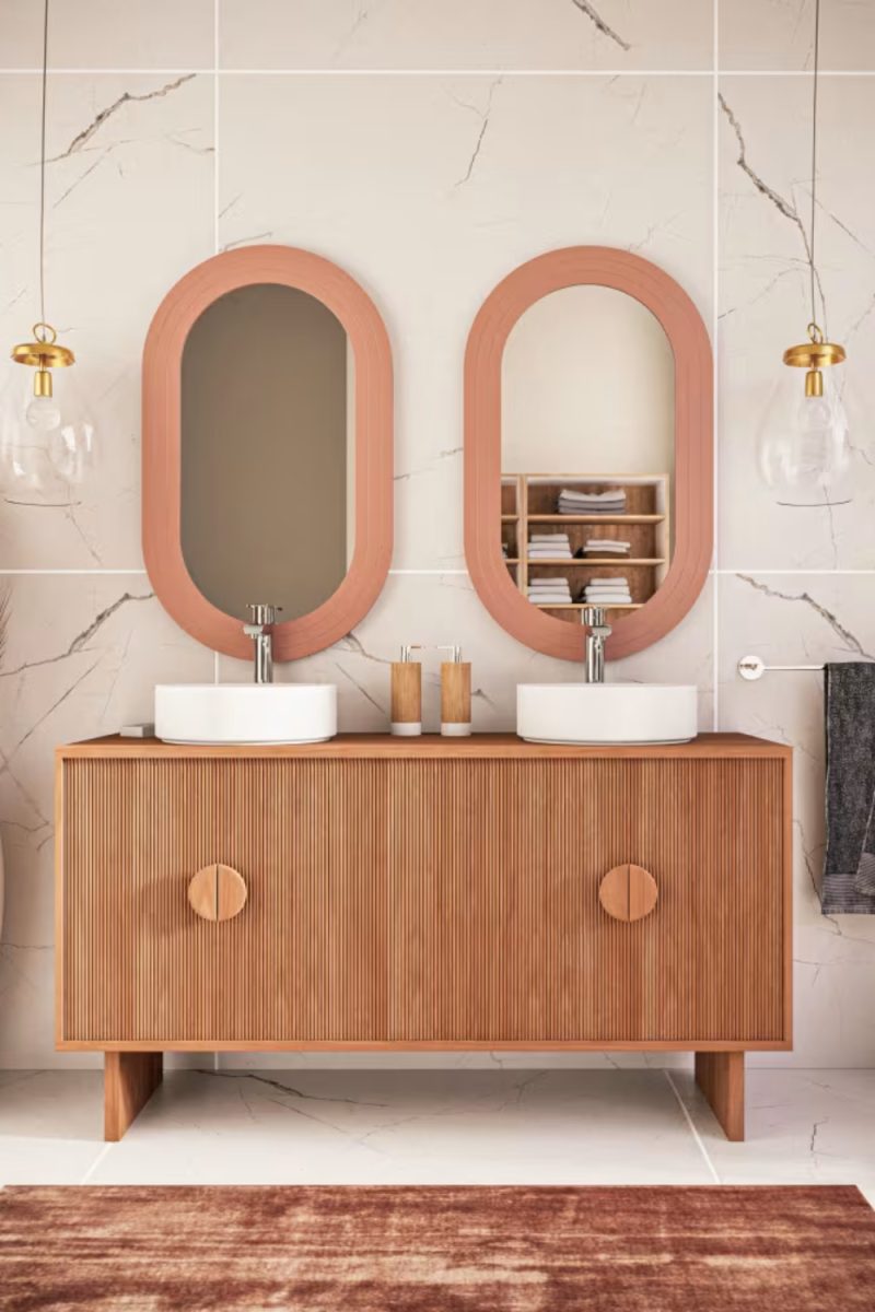 Un meuble-lavabo en bois avec deux lavabos ronds blancs est présenté sous deux miroirs ovales encadrés de couleur pêche dans une salle de bain moderne.