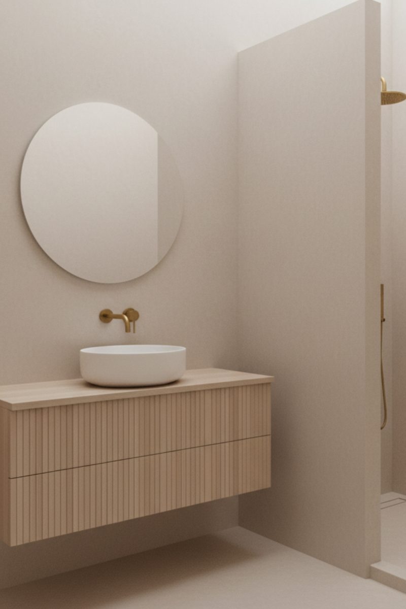 Un lavabo blanc repose sur un meuble en bois clair dans une salle de bain beige minimaliste. Un miroir rond est au-dessus, avec une douche ouverte visible sur le côté droit.