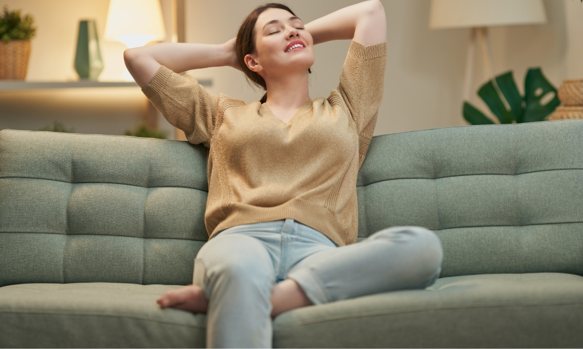 Une femme se détend sur un canapé vert clair, les mains derrière la tête et les yeux fermés, dans un salon lumineux et confortable.