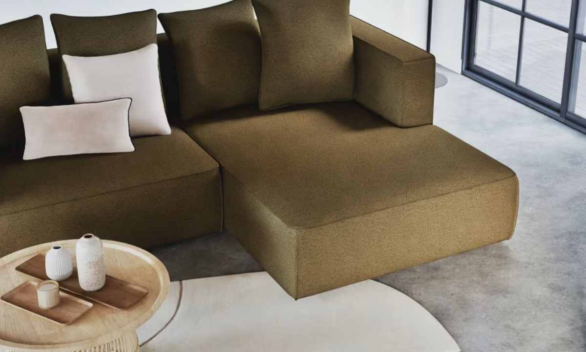 Un canapé d'angle texturé couleur olive présente des coussins assortis et clairs, situé à côté d'une table basse ronde dans un intérieur moderne et minimaliste.