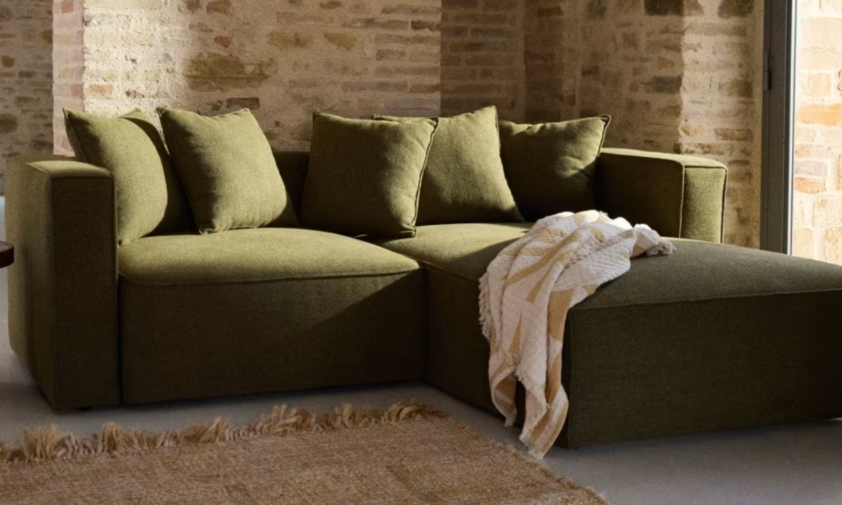 Un canapé d'angle vert olive est placé devant un mur de briques. Un tapis en fibres naturelles est au premier plan et une couverture est posée sur la méridienne.