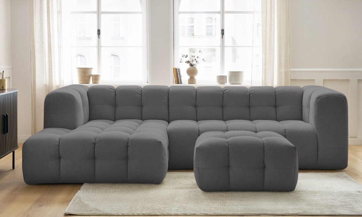 Un grand canapé sectionnel gris est placé sur un tapis clair devant une grande fenêtre lumineuse dans un intérieur minimaliste et moderne.