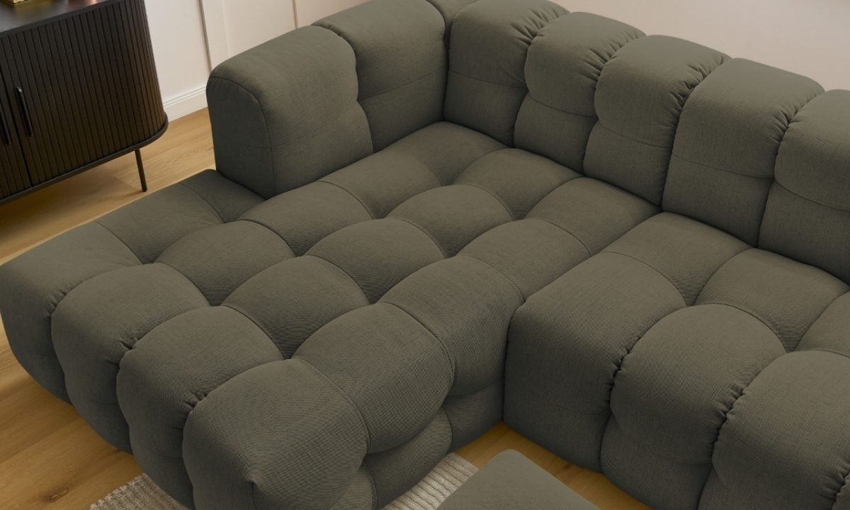 Un canapé modulaire en tissu gris-vert capitonné est disposé en angle sur un plancher en bois clair, avec un meuble sombre à proximité.