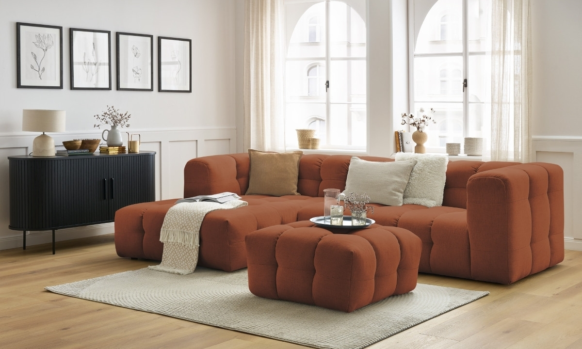 Un canapé modulaire en tissu orange occupe le centre, entouré d'un tapis clair, d'une commode noire et de fenêtres lumineuses dans un intérieur moderne et chaleureux.