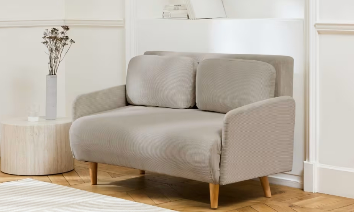 Un fauteuil beige en velours côtelé repose sur un parquet. À côté, une table ronde en bois accueille un vase et un verre, le tout dans un intérieur minimaliste.