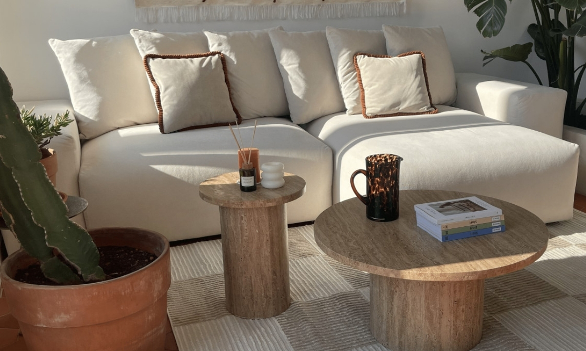 Un salon minimaliste est éclairé par la lumière naturelle. Un canapé blanc confortable est agrémenté de coussins, et des tables basses en pierre soutiennent des objets décoratifs.