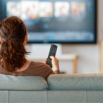 Voici une description objective de l'image : Une femme assise sur un canapé regarde la télévision, une télécommande à la main. L'arrière-plan est flou, avec des meubles et des étagères.