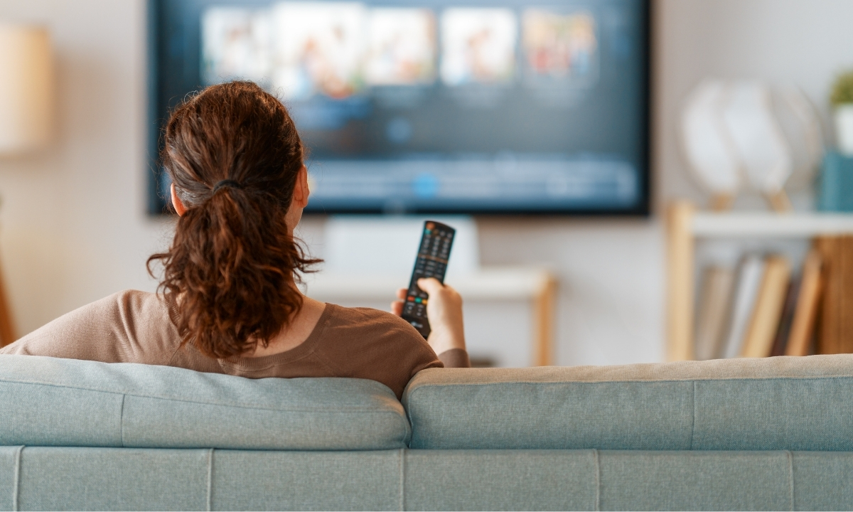 Voici une description objective de l'image : Une femme assise sur un canapé regarde la télévision, une télécommande à la main. L'arrière-plan est flou, avec des meubles et des étagères.