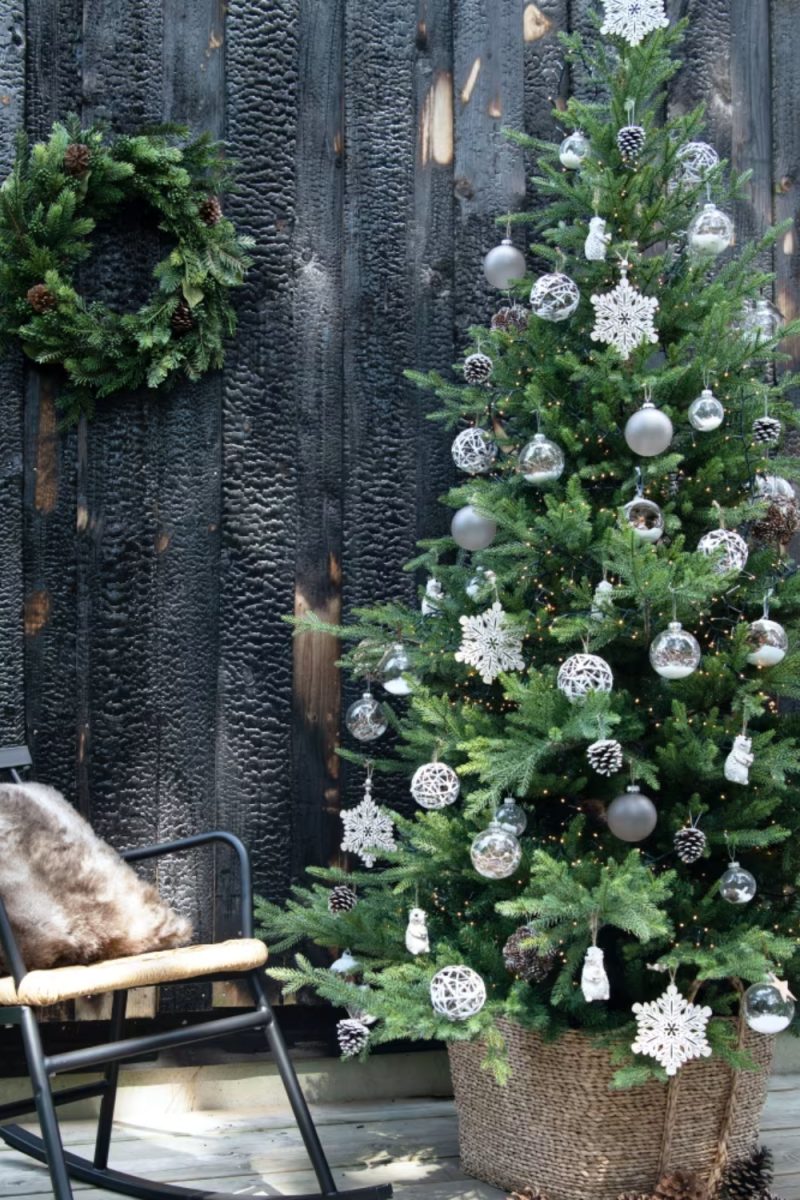 Un arbre de Noël décoré est installé à l'extérieur, à côté d'une chaise avec un coussin en fourrure, devant un mur en bois sombre et une couronne.