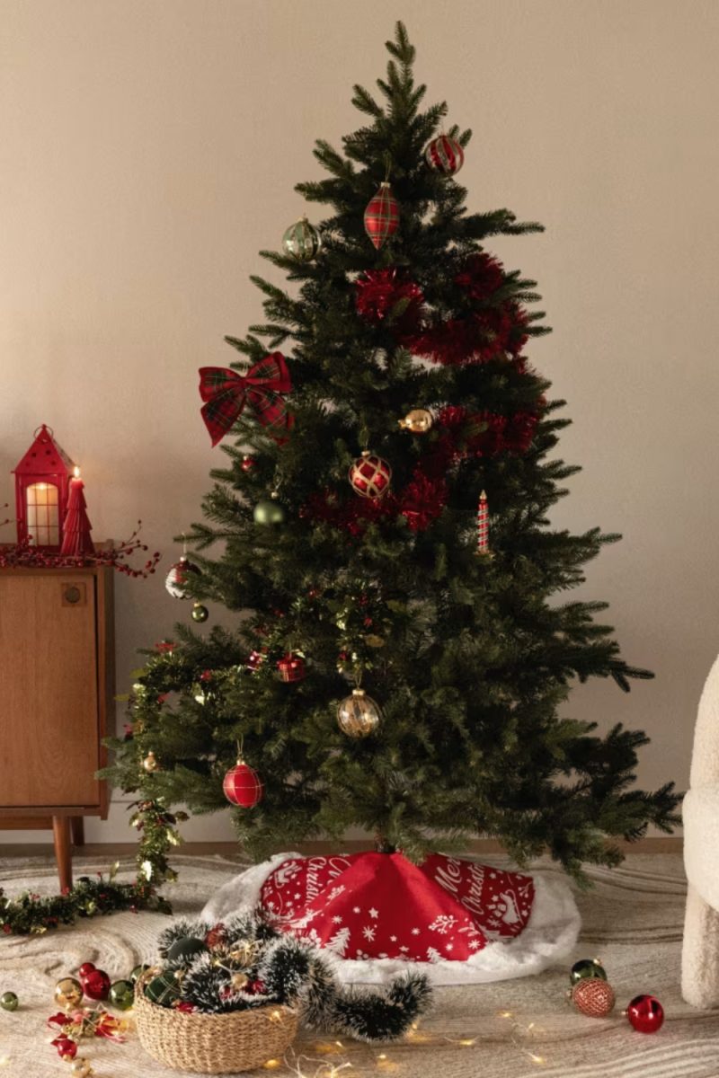 Un sapin de Noël décoré trône dans une pièce, orné de guirlandes rouges, de boules colorées et d'un nœud à carreaux. Un tapis de sapin rouge et blanc est à sa base.