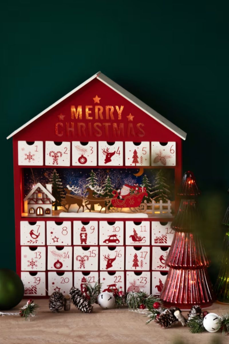 Un calendrier de l'Avent en forme de maison, avec l'inscription "MERRY CHRISTMAS", est entouré de décorations de Noël sur une surface en bois devant un fond vert foncé.
