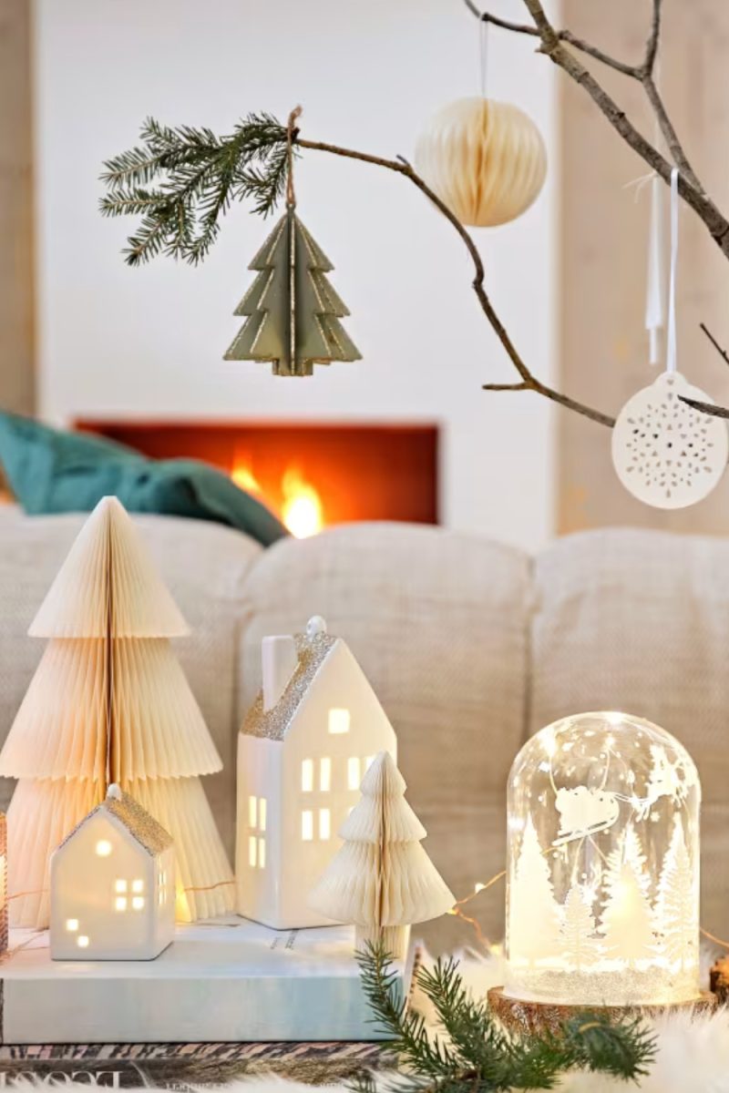 Décorations de Noël lumineuses et élégantes, incluant des maisons miniatures et des arbres en papier, posées sur un livre, avec une cheminée en arrière-plan.