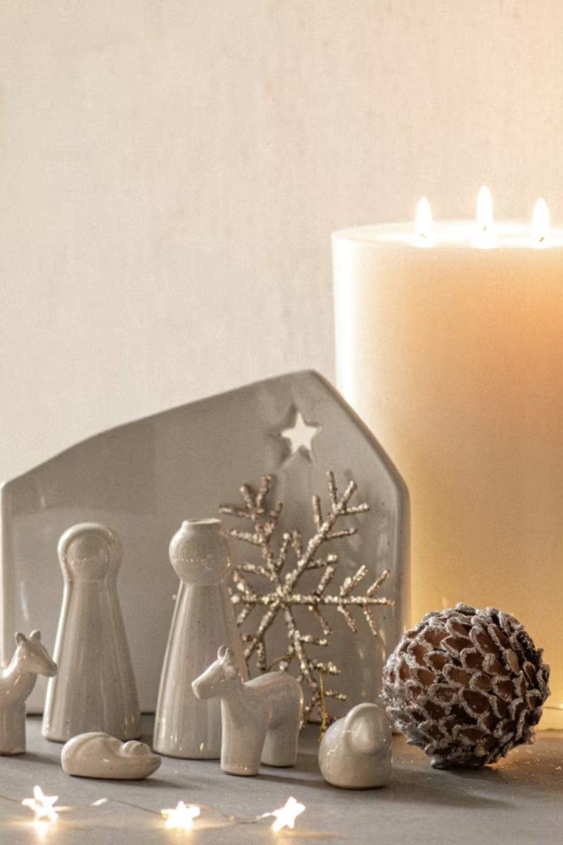 Un arrangement de Noël monochrome présente une crèche minimaliste, une bougie allumée et une pomme de pin décorative sur une surface grise, sous une douce lumière.