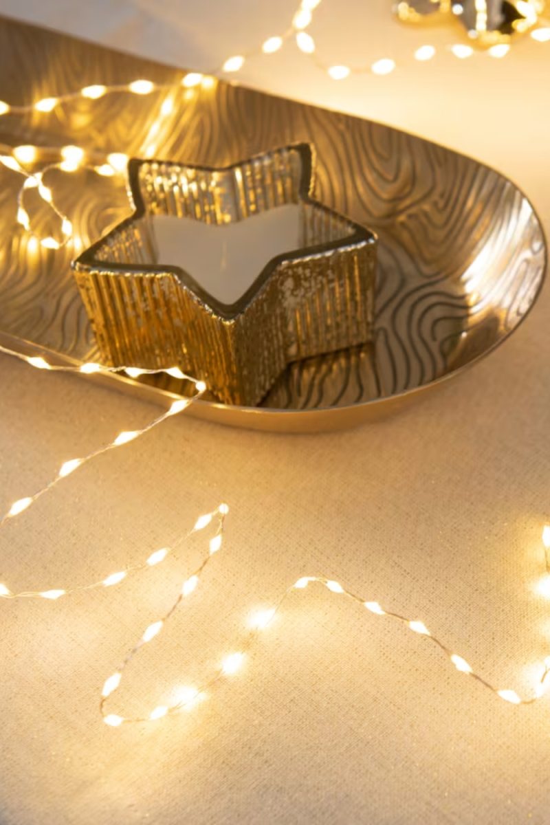 Une bougie en forme d'étoile dorée repose sur un plateau texturé doré. Des guirlandes lumineuses blanches scintillent autour, créant une ambiance chaleureuse et festive.