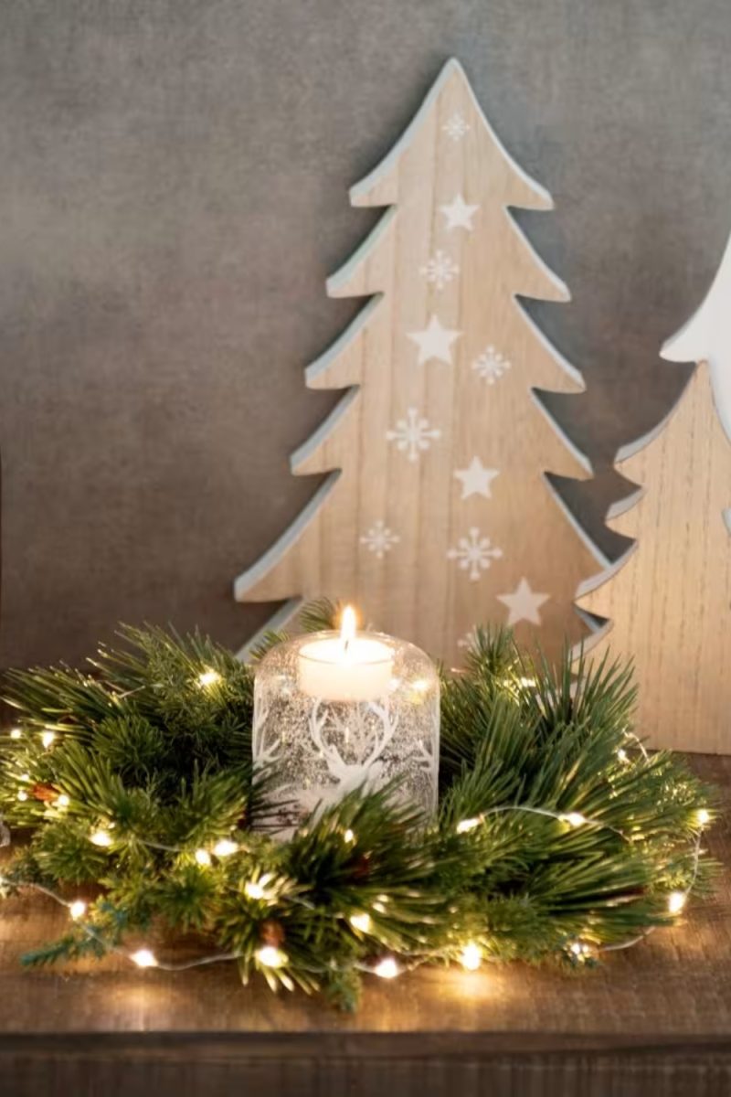 Un arrangement festif de Noël avec une bougie dans un photophore à motif de cerf, entourée de verdure et de guirlandes lumineuses, devant des arbres décoratifs en bois.