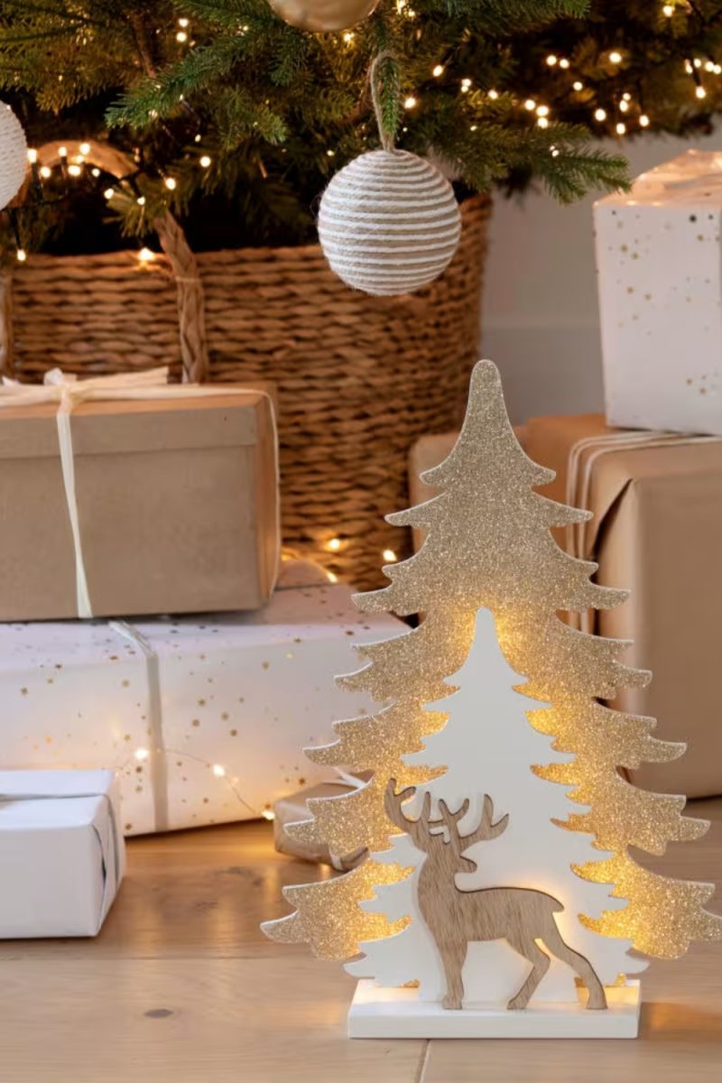 Décoration de Noël : un arbre doré illuminé avec un renne en bois, placé devant des cadeaux et un arbre décoré de lumières.