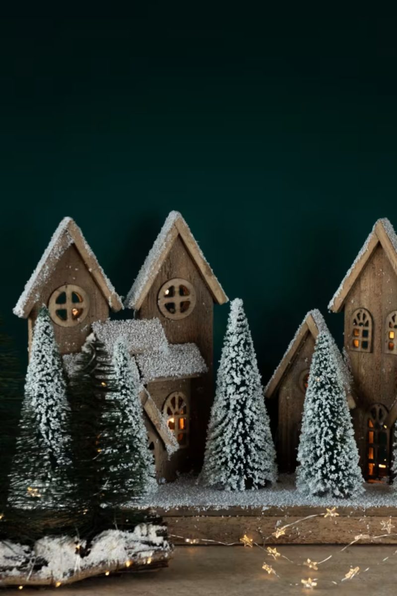 Des maisons en bois enneigées et des sapins sont illuminés par des guirlandes lumineuses dans un décor festif vert foncé.