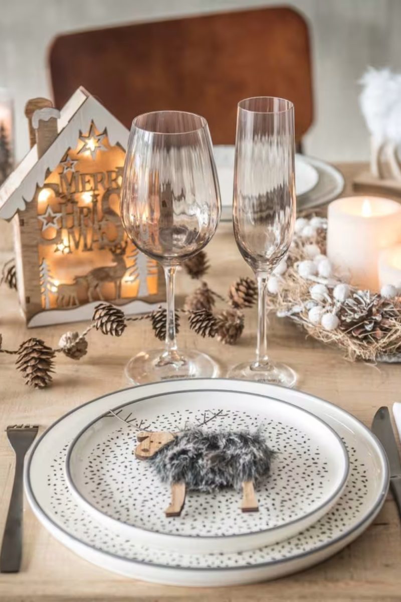 Voici une table dressée pour Noël. Une assiette décorée avec un ornement d'agneau repose sur une table en bois avec des verres, une maison lumineuse et des décorations naturelles. "MERRY CHRISTMAS" est inscrit sur la maison.
