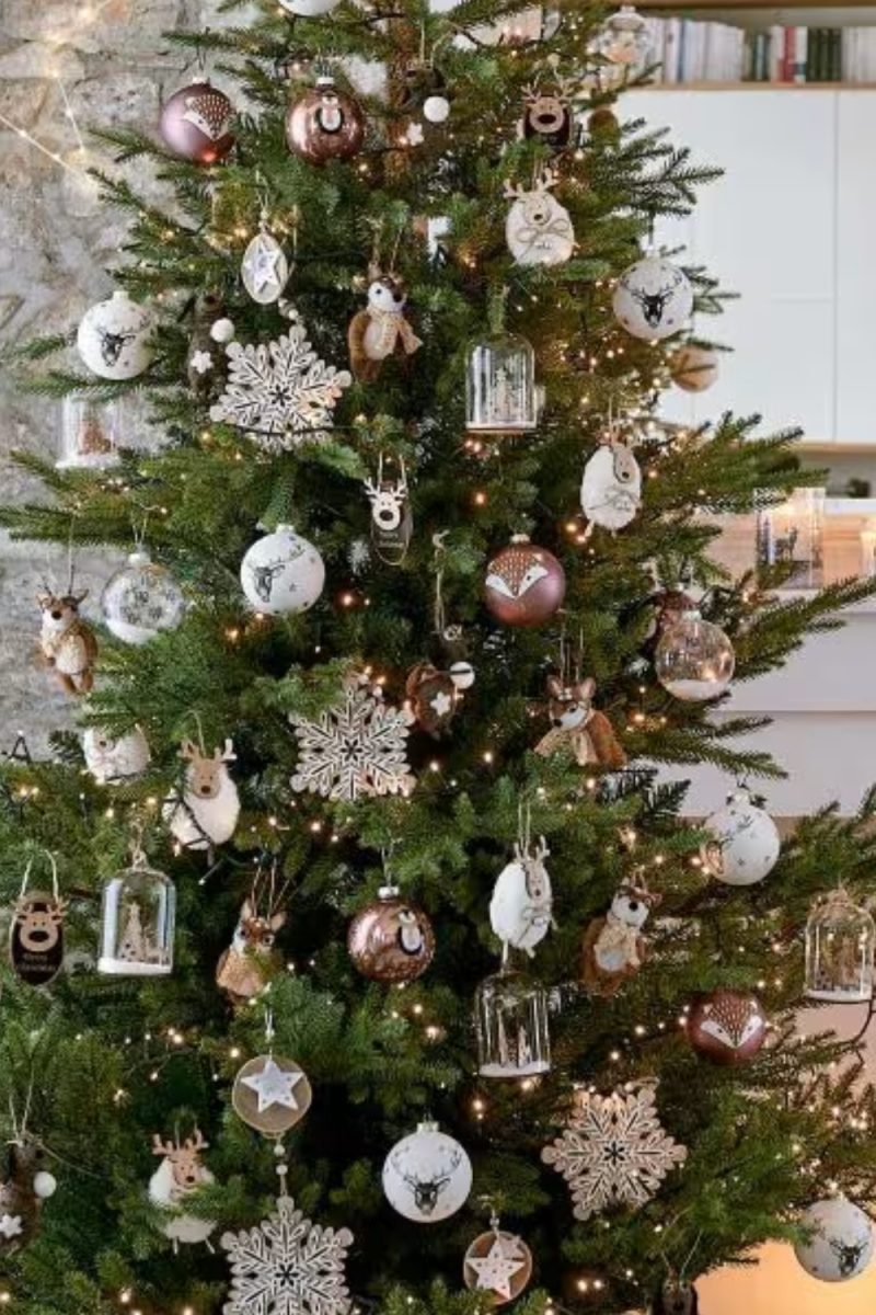 Un sapin de Noël dense est décoré avec des boules, des flocons de neige et des figurines d'animaux, illuminé par des guirlandes lumineuses dans un intérieur chaleureux.