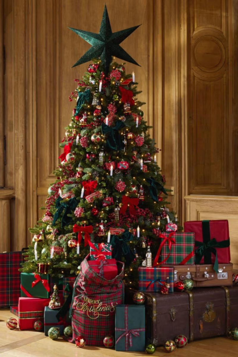 Un arbre de Noël décoré trône devant un mur boisé. Des cadeaux emballés sont disposés à son pied, avec un sac "Merry Christmas" tartan.