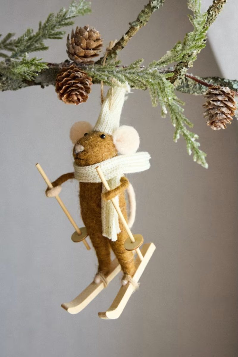 Une décoration de Noël représentant une souris skieuse est suspendue à une branche de conifère décorée de pommes de pin, sur un fond uni.