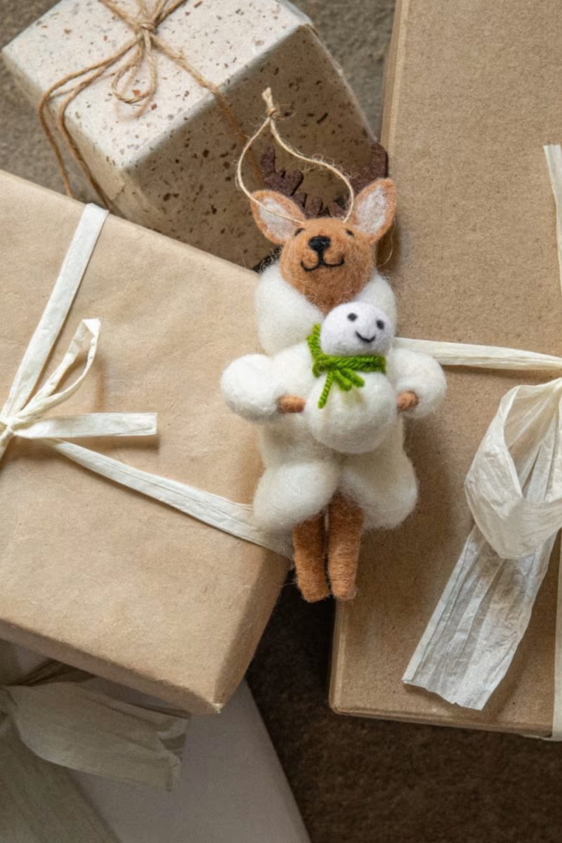 Voici une description de l'image : Un ornement de renne en feutre tient un bonhomme de neige, reposant sur des cadeaux emballés dans du papier brun et beige, noués avec de la ficelle et du ruban.