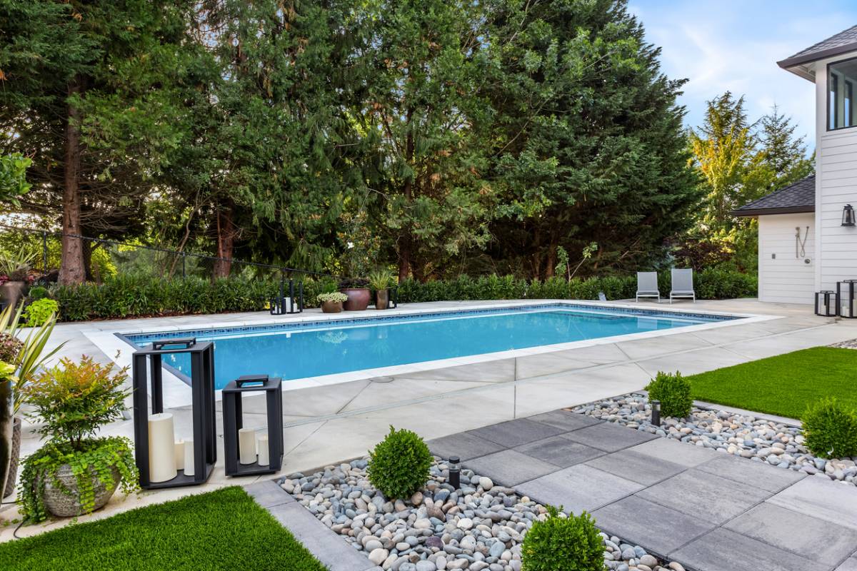 Une piscine bleue rectangulaire est entourée d'une terrasse en béton, de verdure luxuriante et de quelques éléments décoratifs comme des lanternes près d'un carré d'herbe verte et galets.