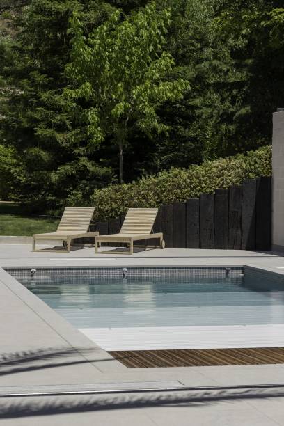 Voici une description de l'image: Deux chaises longues en bois sont au bord d'une piscine partiellement recouverte, sur une terrasse pavée, avec un jardin boisé en arrière-plan.