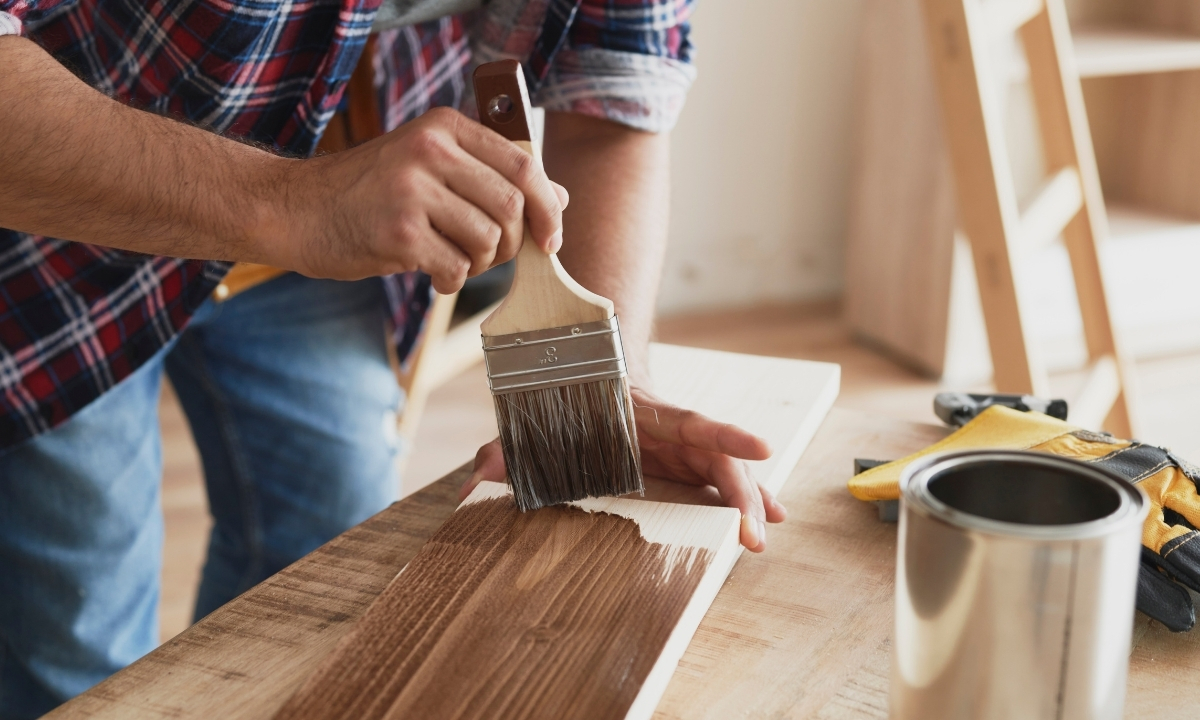 Un homme en chemise à carreaux vernit une planche de bois avec un pinceau dans un atelier de menuiserie, avec un pot de vernis et des gants à proximité.